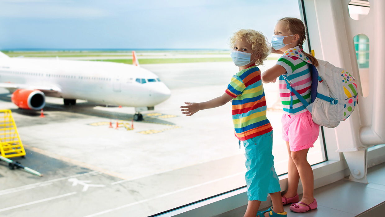 Two children wearing masks looking out a window at an airport.