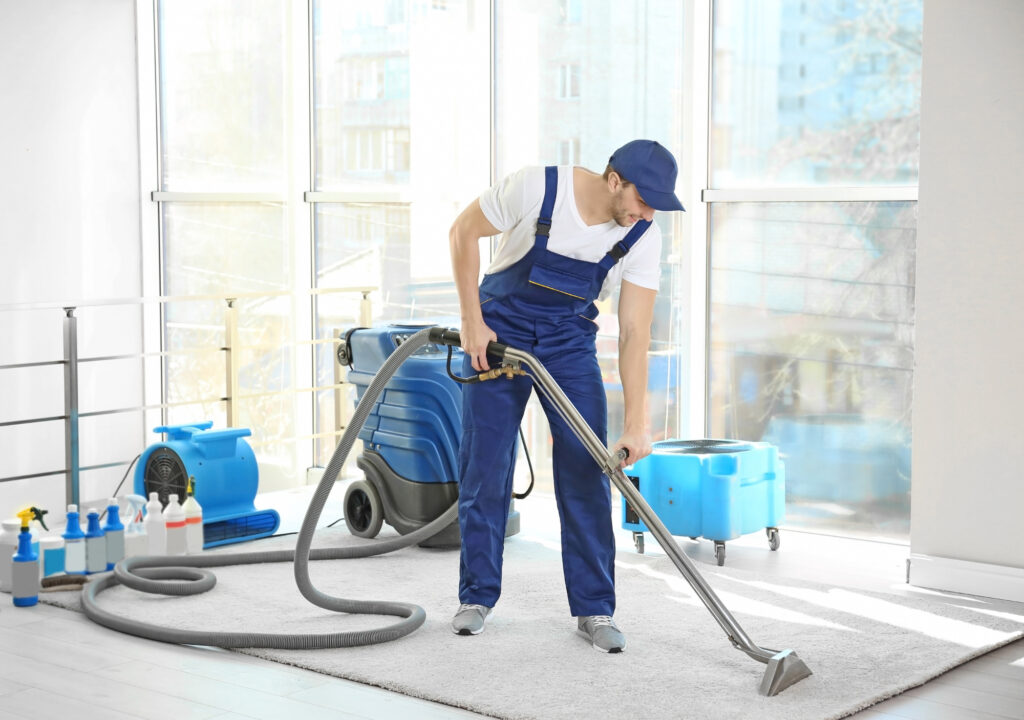 A man in blue overalls is vacuuming the floor.